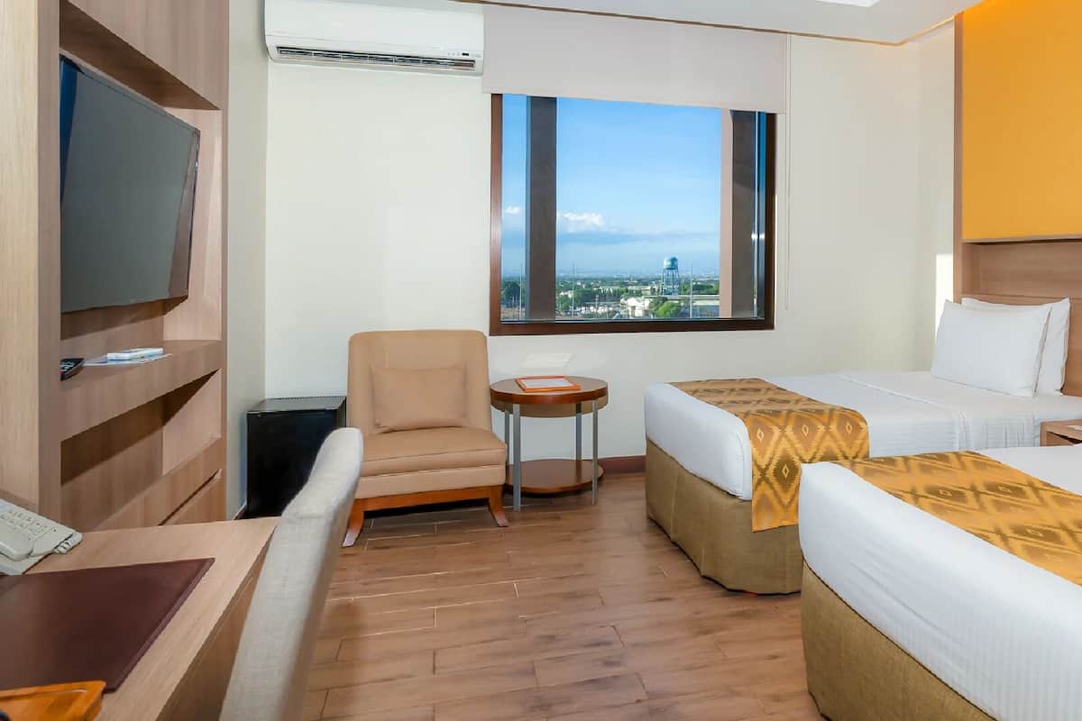 Habitación de hotel con dos camas, televisor, una silla, una mesita y ventana con vista a la ciudad.