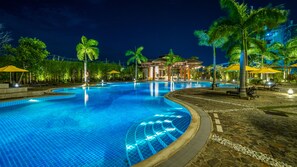 Una piscina iluminada de noche con agua cristalina, rodeada de palmeras y zonas de estar al aire libre.