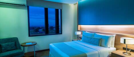 A hotel room with a large bed, a nightstand, a lamp, a chair, and a window with a city view.