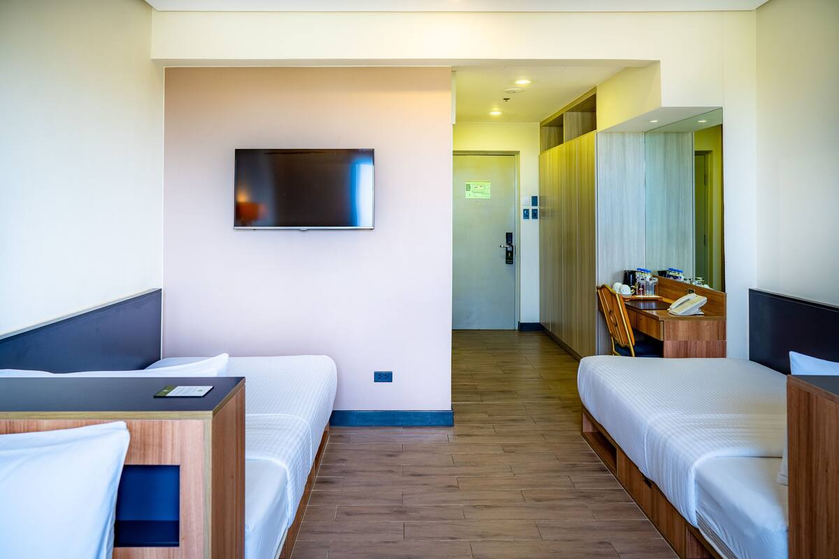 Habitación de hotel con dos camas, un televisor montado en la pared, un escritorio con una silla y un baño visible a través de una puerta abierta.