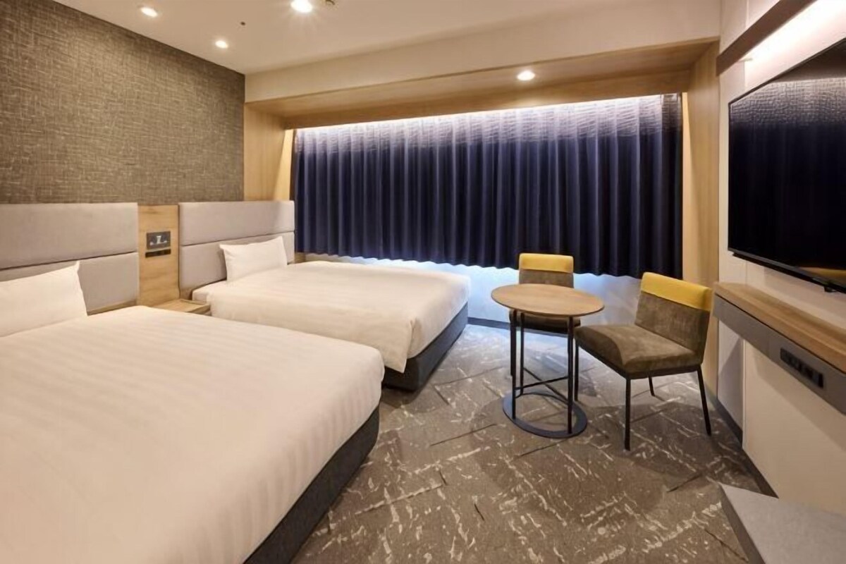 A hotel room with two beds, a small table, a chair, a TV, and a large window with curtains.
