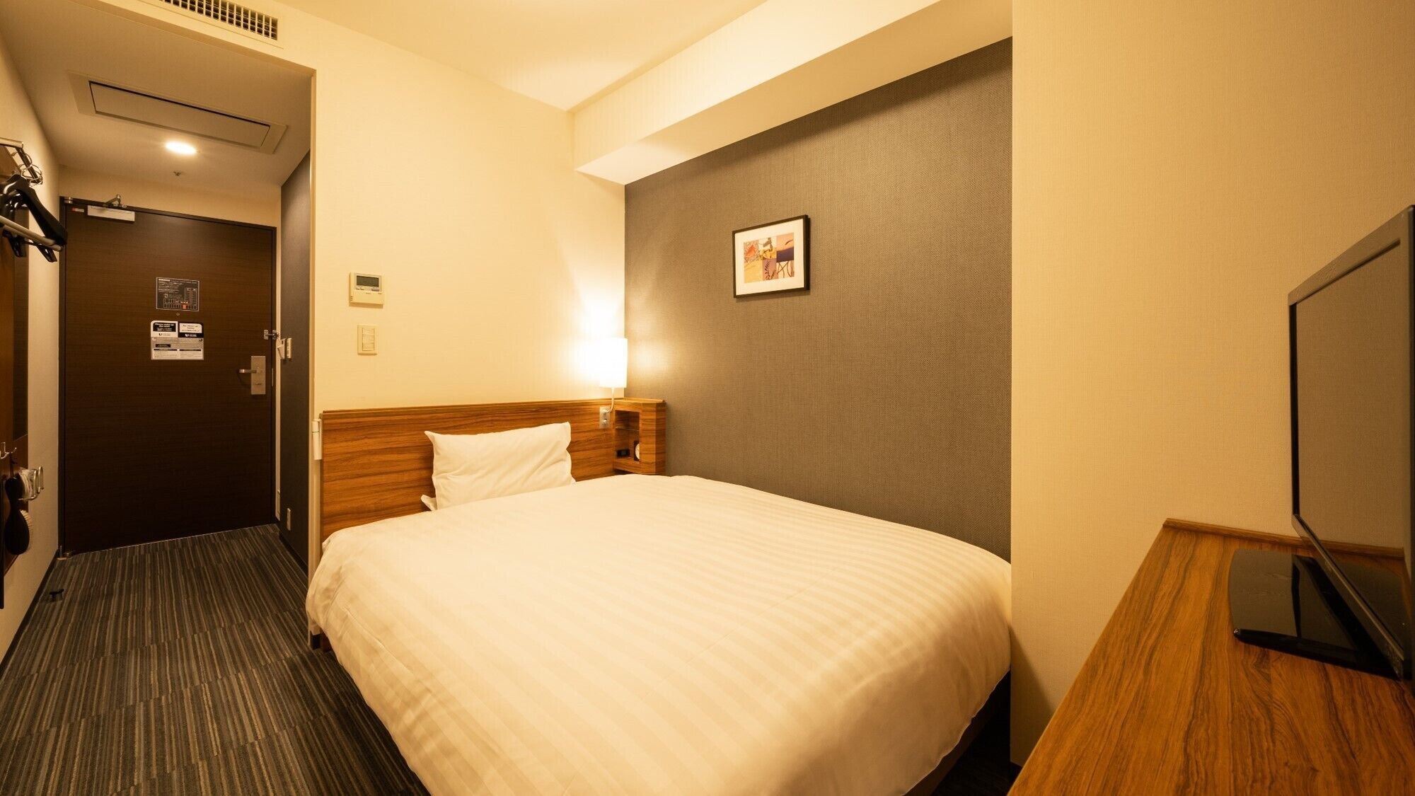 A hotel room with a bed, a bedside table, a wall-mounted TV, and a door leading to a hallway.
