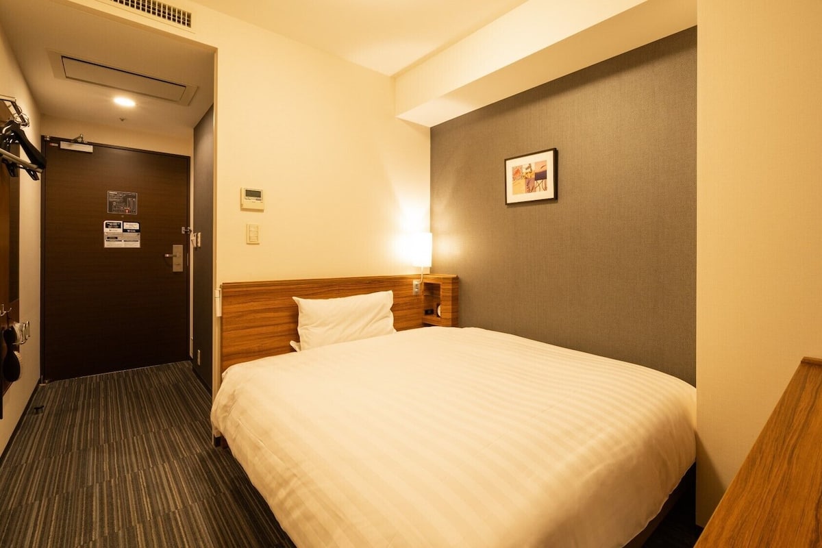 A hotel room with a bed, a bedside table, a wall-mounted TV, and a door leading to a hallway.