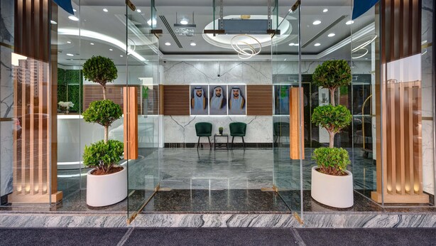 Un área de recepción moderna con paredes de vidrio, dos plantas en macetas, una zona de estar con sillas y un suelo de mármol.