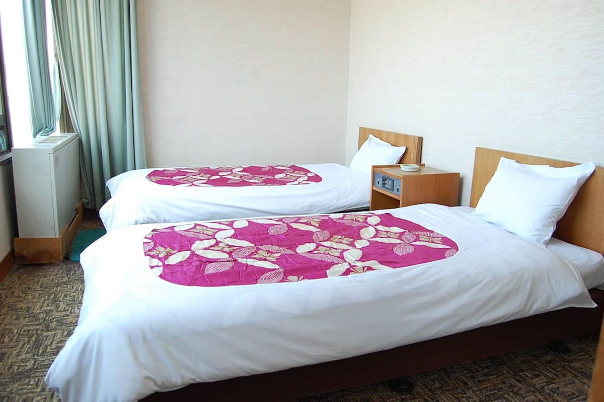 Habitación de hotel con dos camas, cada una con cubrecama floral y ropa de cama blanca. Tiene cabecera de madera, mesita de noche con lámpara y ventana con cortinas.