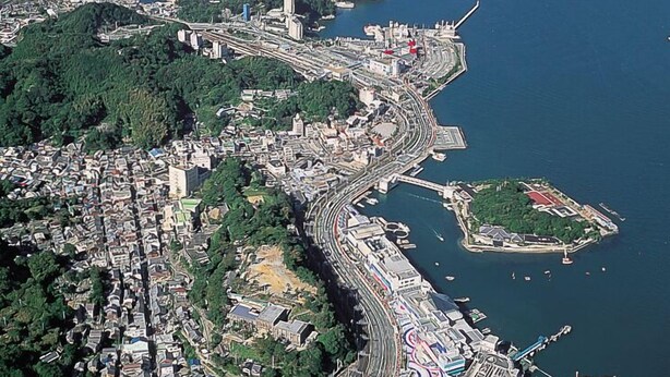 An aerial view of a coastal city with a dense urban area, a prominent curved road, and several islands in the background. - Kinpokan (Toba)