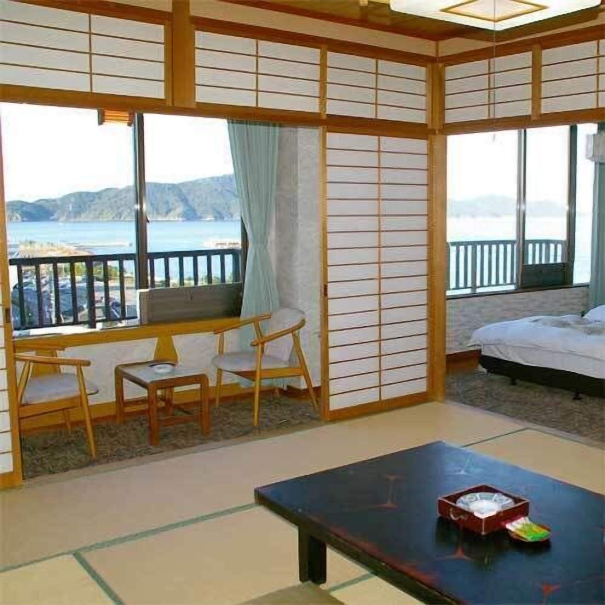 A traditional Japanese-style room with tatami flooring, a low table with chairs, a bed, and large windows with a view of the sea.