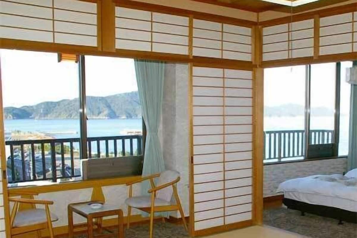 Una habitación tradicional japonesa con suelo de tatami, una mesa baja con sillas, una cama y grandes ventanas con vista al mar.