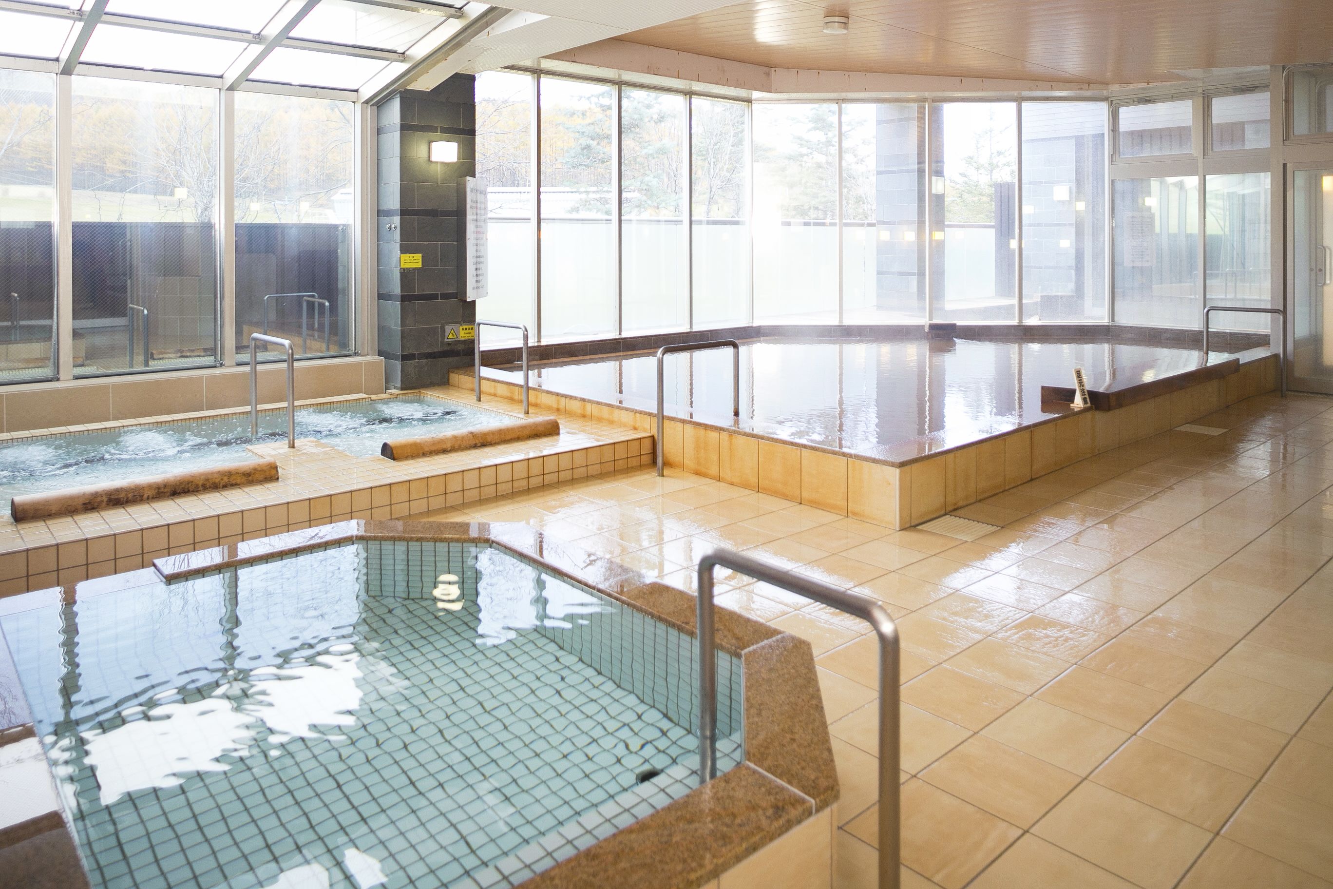A large indoor swimming pool with clear glass walls, tiled floors, and a wooden deck area.