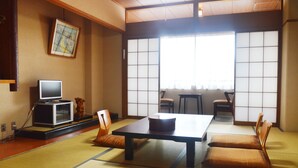 Una habitación japonesa tradicional con suelo de tatami, una mesa baja, cojines, un televisor y puertas correderas.