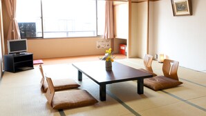 Una habitación tradicional japonesa con suelo de tatami, una mesa baja con cojines, un televisor y un amplio ventanal con cortinas.
