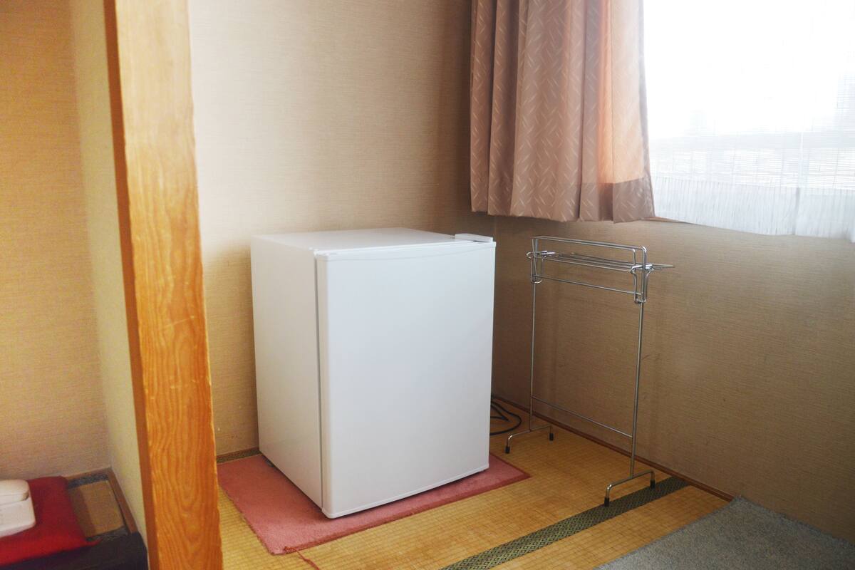 A compact room with a white refrigerator, a metal drying rack, and a window with curtains.