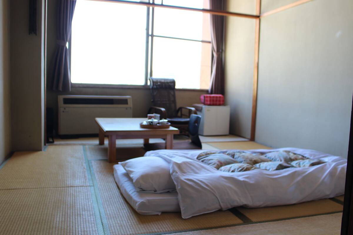 Une pièce de style japonais traditionnel, avec un sol recouvert de tatamis, un futon servant de lit, une petite table avec un service à thé et une fenêtre avec des rideaux.