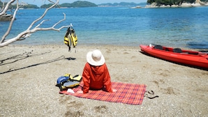 Uma pessoa sentada na praia, com um caiaque vermelho e coletes salva-vidas por perto.