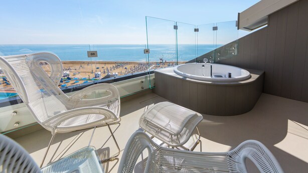 Un balcón con sillones de mimbre, un jacuzzi y baranda de vidrio con vista a la playa.