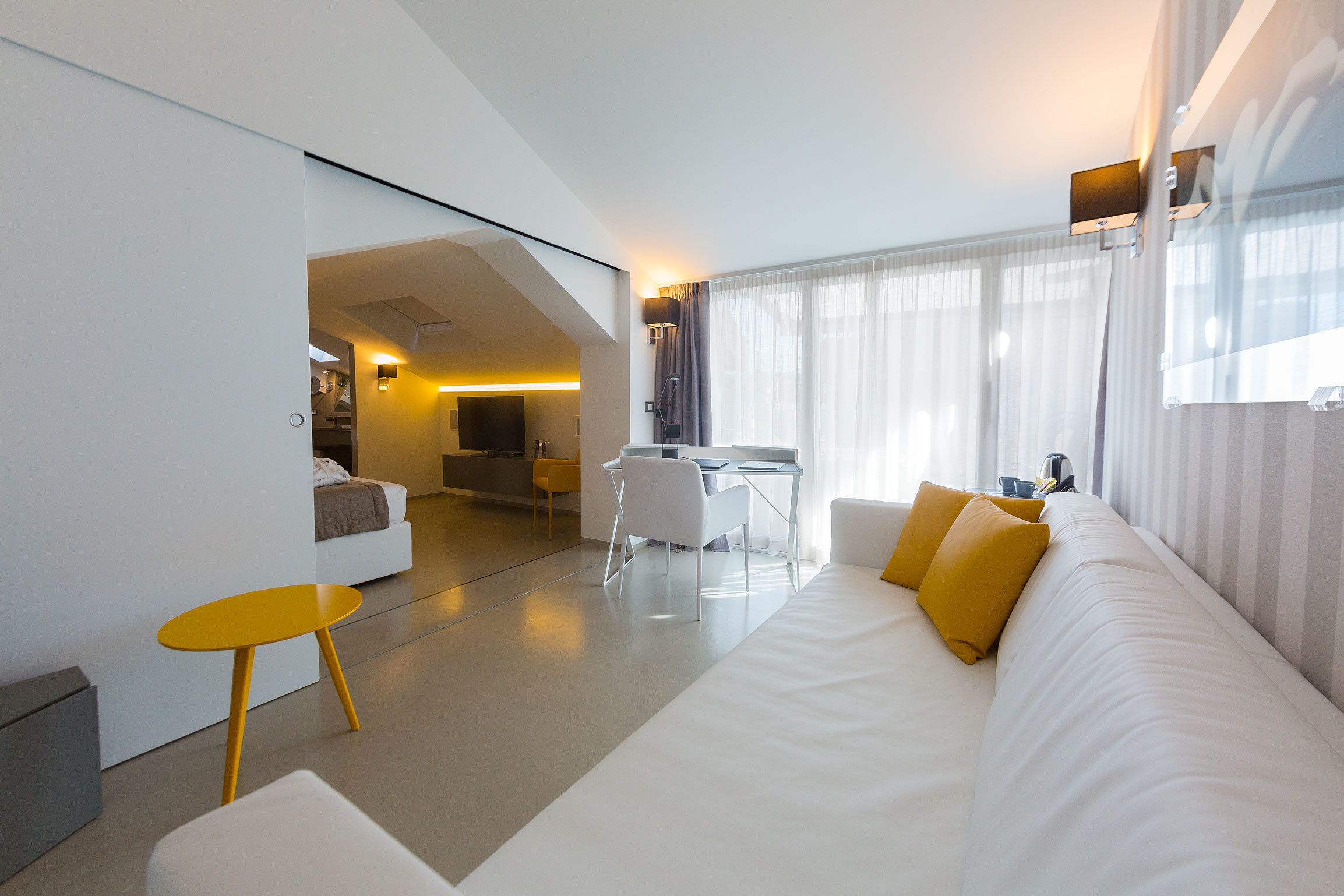 Une chambre d’hôtel moderne avec un canapé blanc, une petite table jaune et un coin repas comprenant une chaise blanche.