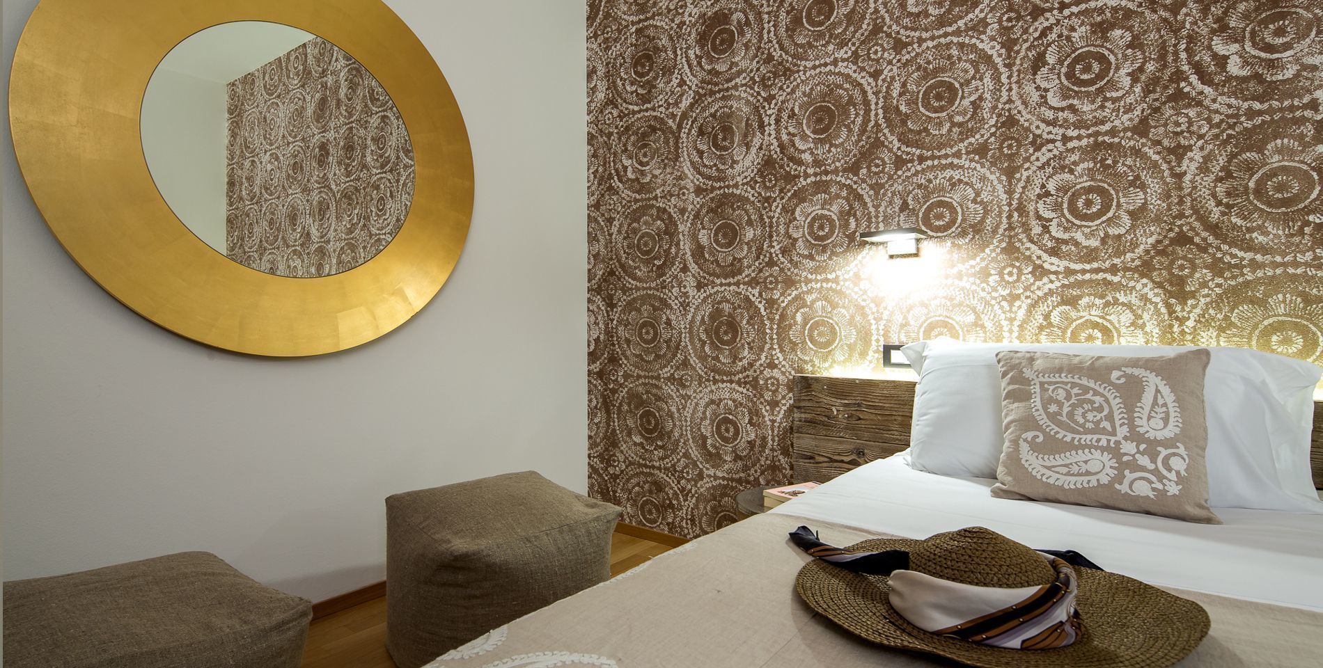 A hotel room with a round mirror, patterned wallpaper, a bed with pillows, a nightstand, and a woven hat on the bed.