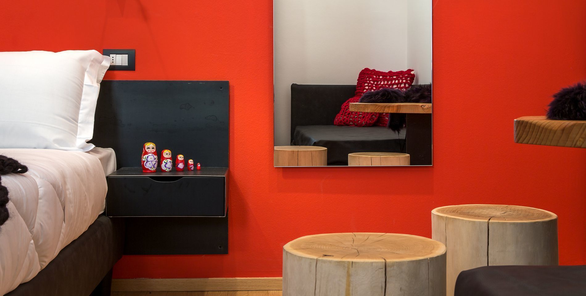 Une chambre avec un mur rouge, un lit recouvert d’une literie blanche, une table de chevet avec un miroir et une petite collection de poupées russes gigognes.