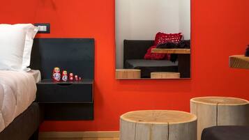Une chambre avec un mur rouge, un lit recouvert d’une literie blanche, une table de chevet avec un miroir et une petite collection de poupées russes gigognes.