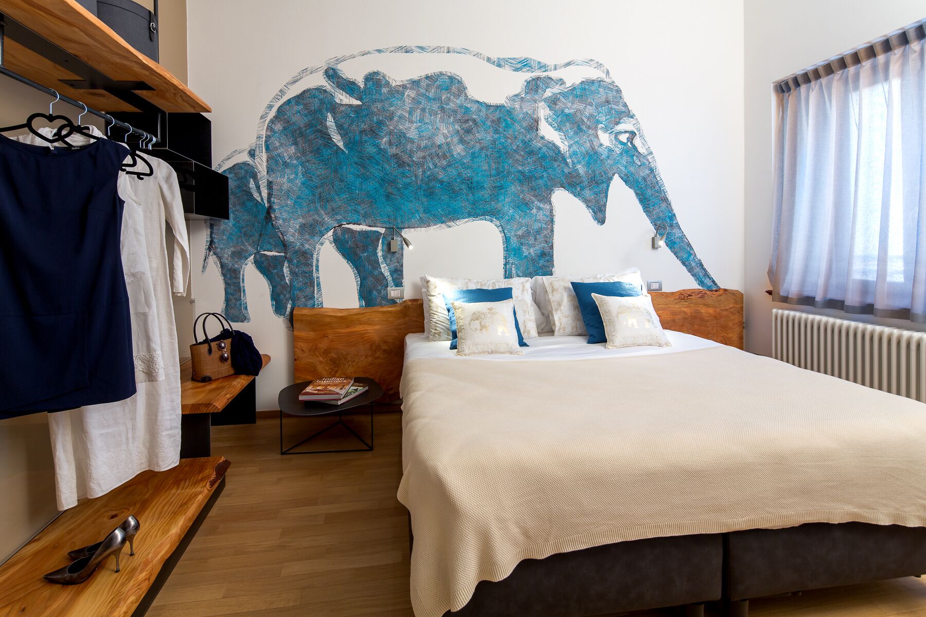 Un dormitorio con un amplio mural de un elefante azul, una cama con ropa de cama blanca y azul, una mesita de noche de madera y una ventana con cortinas de encaje.