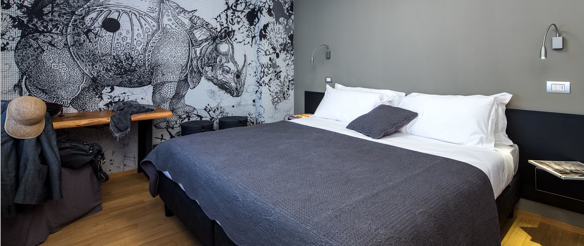 A modern hotel room with a large bed, a desk with a chair, and a wall with a black and white patterned tapestry.