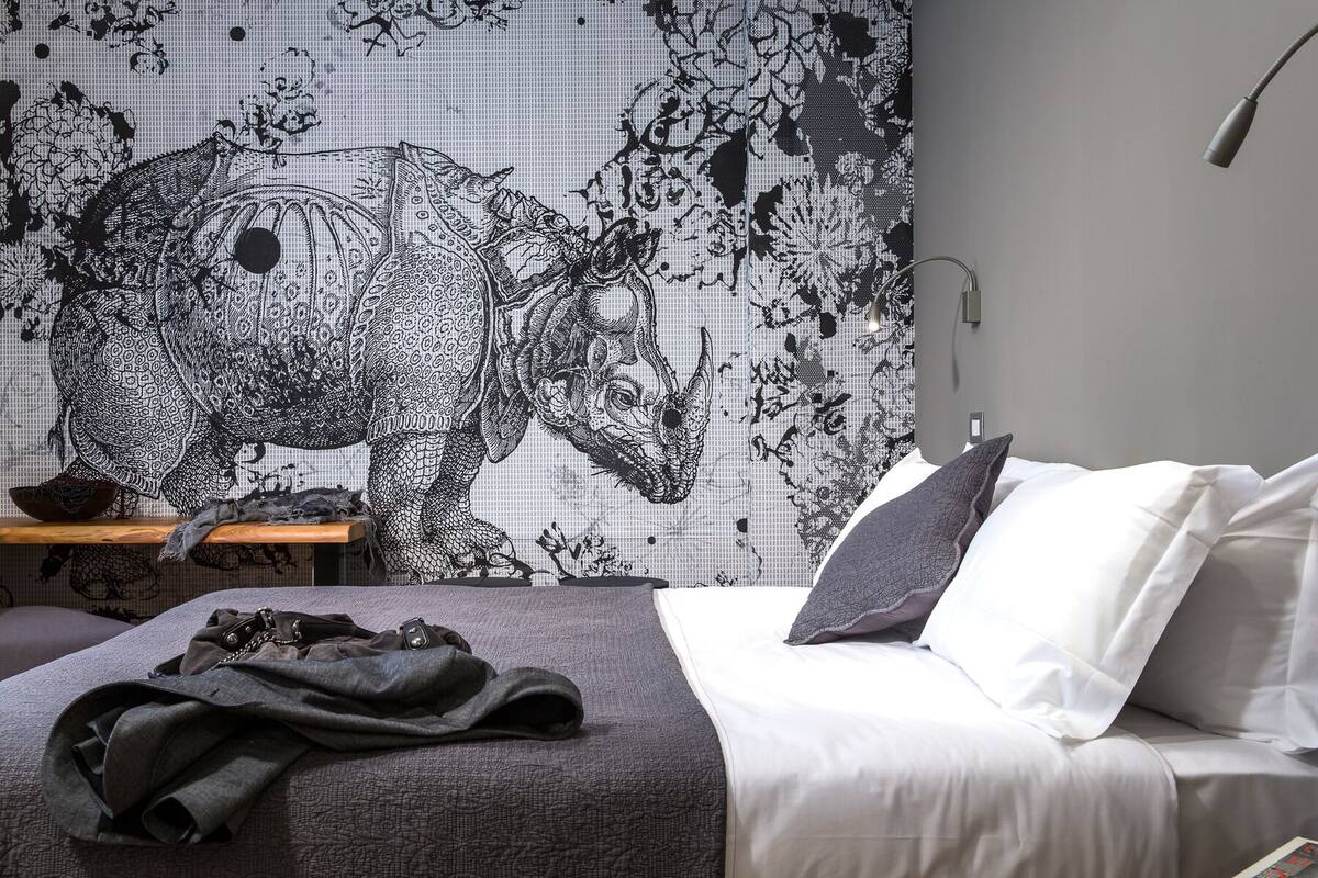 Een slaapkamer met een groot muurschildering van een neushoorn, een bed met wit en grijs beddengoed, en een klein houten tafeltje met een schaal en een tas.