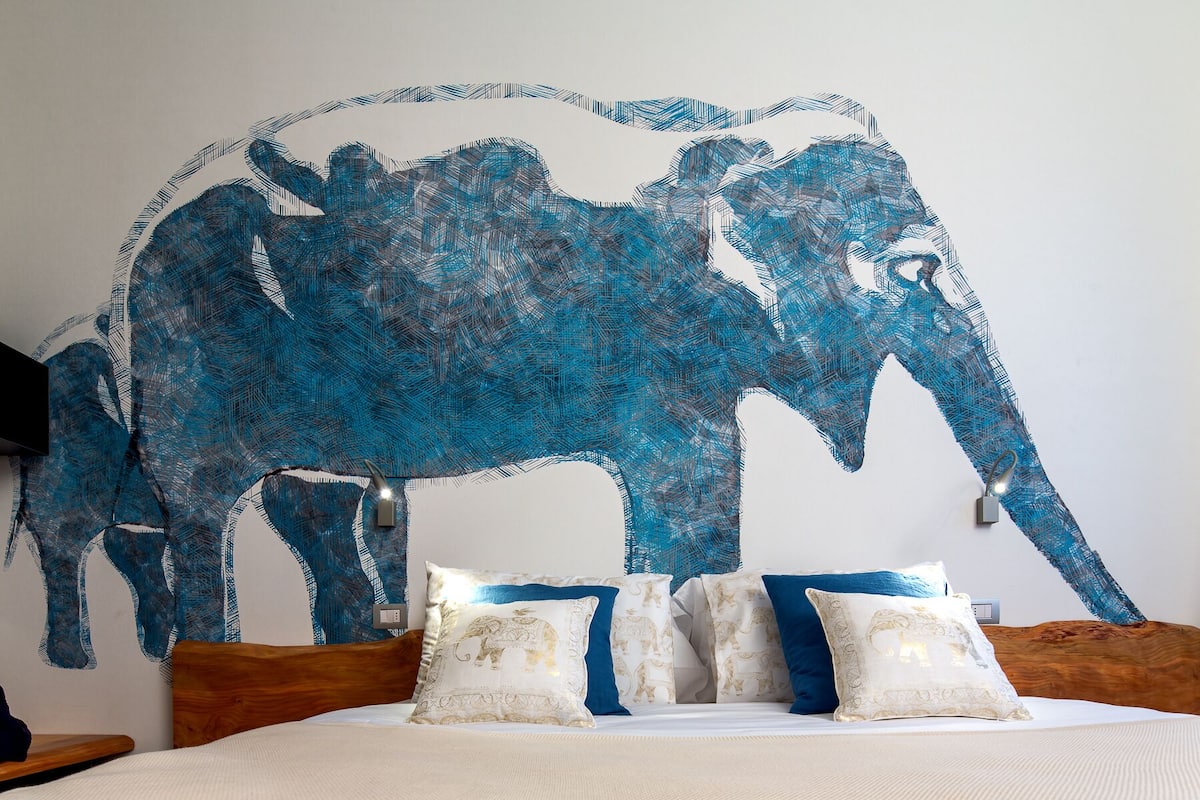 Een slaapkamer met een groot wanddecoratie van een olifant, een houten hoofdbord en een bed met witte beddengoed en blauwe kussens.