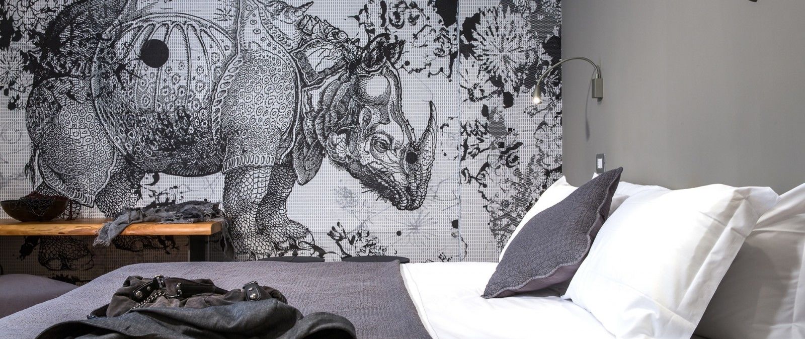 Un dormitorio con un gran tapiz detallado en la pared, una cama con ropa de cama blanca y gris, y una mesita de noche de madera con un cuenco y una cámara.