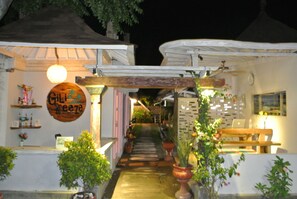 A narrow pathway leading to a building with a wooden sign reading "Gili Freeze." There are potted plants along the path, and a bench is visible in the background.