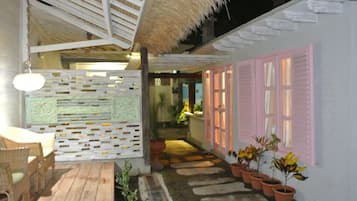 Eine Holzterrasse mit Korbmöbeln, einem Reetdach und Blumentöpfen entlang eines Weges zu einem Gebäude mit rosafarbenen Fensterläden.