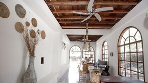 Ein Flur mit Deckenventilator, einem Holztisch, Stühlen, einer Vase mit getrockneten Zweigen und einem Schreibtisch mit Computer.