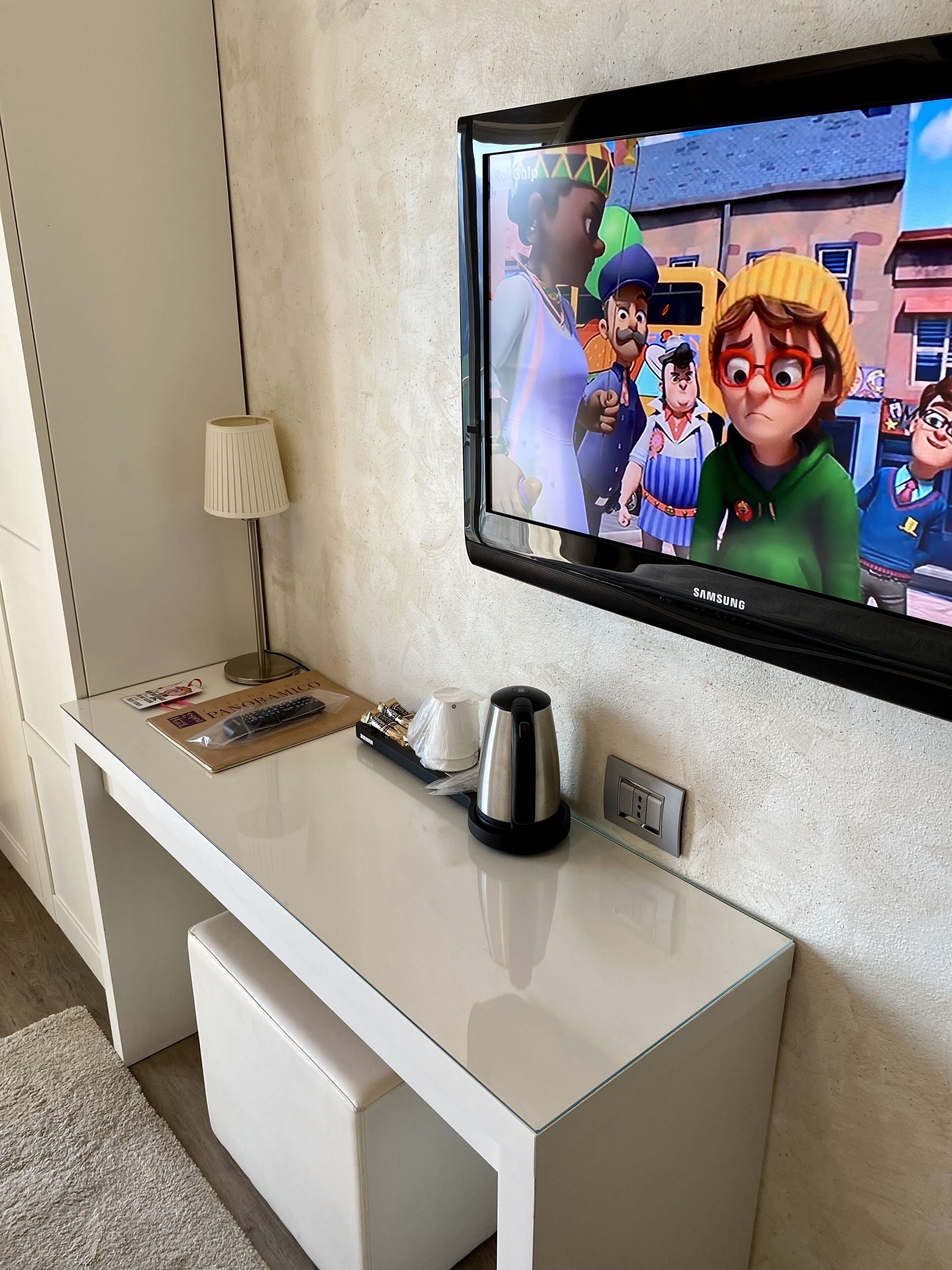 A modern hotel room with a flat-screen TV mounted on the wall, a white desk with a lamp, a kettle, and a remote control.