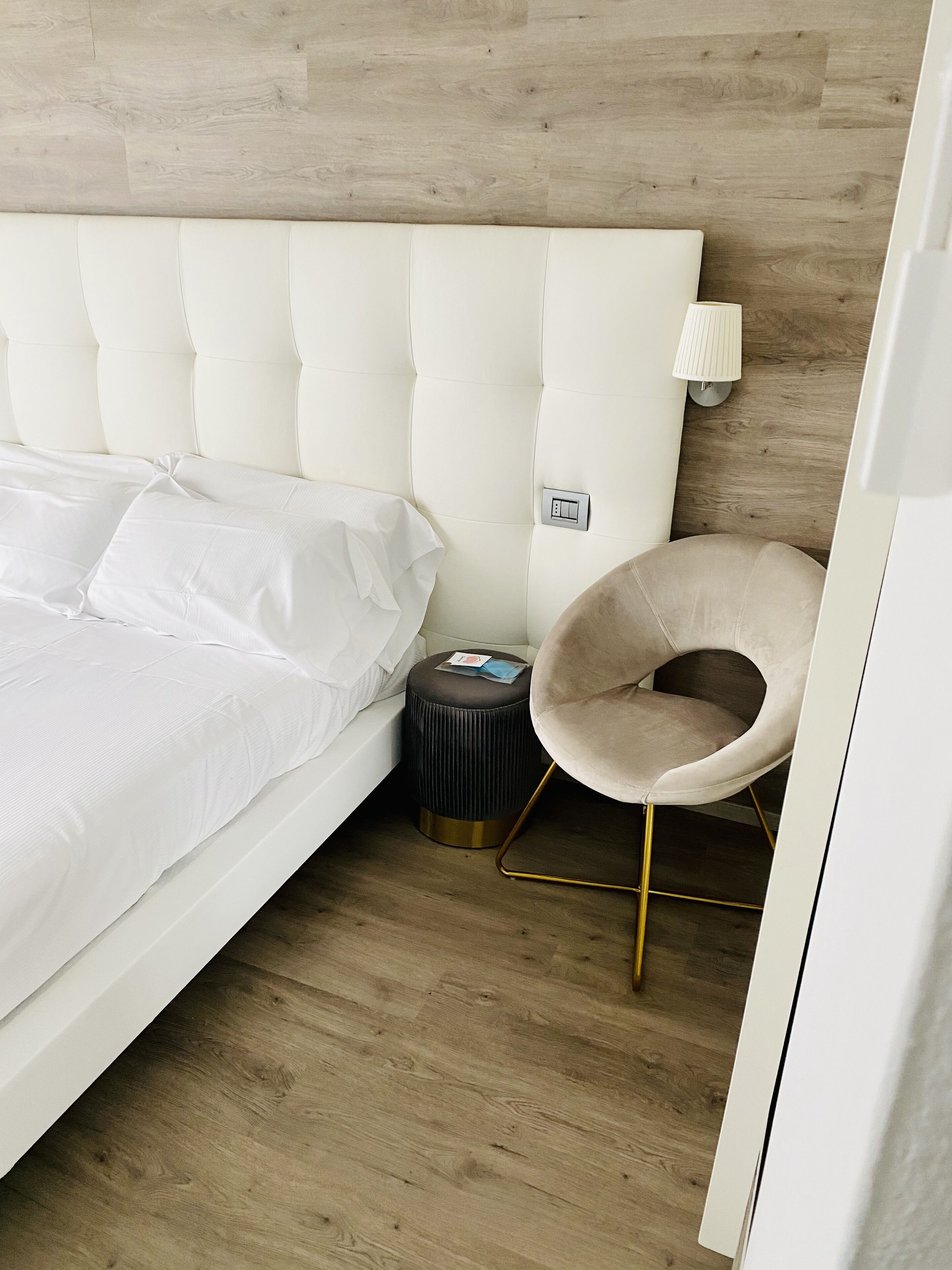 A neatly made bed with white linens, a round ottoman with a lamp, and a chair with a gold frame.
