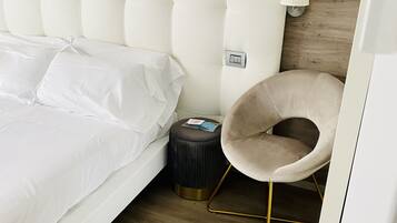 A neatly made bed with white linens, a round ottoman with a lamp, and a chair with a gold frame.