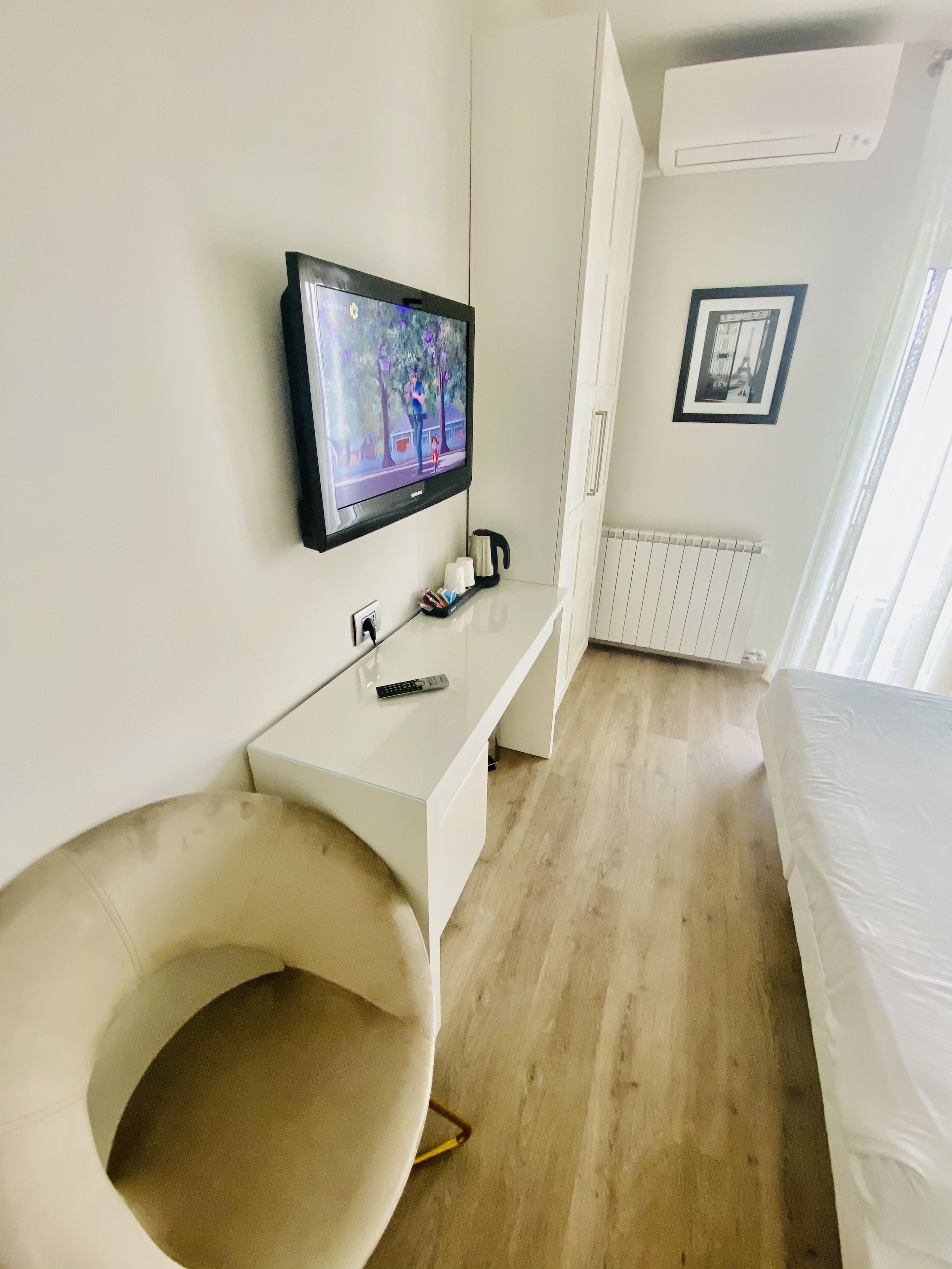 Una habitación de hotel moderna con una televisión de pantalla plana montada en la pared, un escritorio blanco con un control remoto, una silla acolchada, una cama y un radiador.