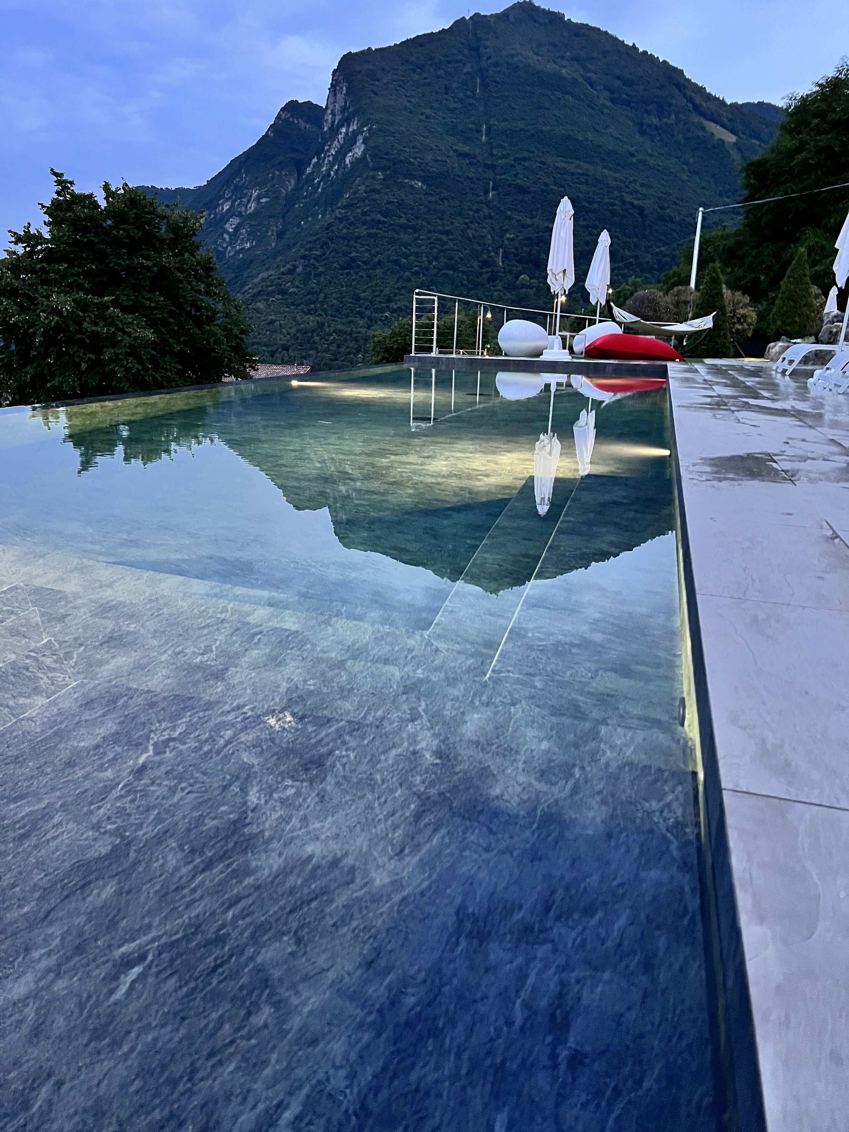 Ein klares Schwimmbecken mit Bergblick, eine Liegewiese mit Sonnenschirmen und einem wolkenlosen Himmel.