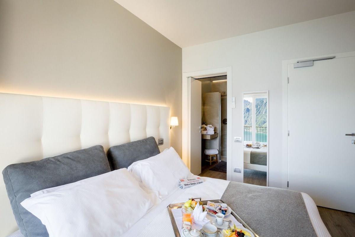 Ein Hotelzimmer mit einem großen Bett, einem Nachttisch mit Frühstücksangebot, einem Badezimmer mit Dusche und Meerblick durch ein Fenster.