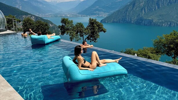 Una piscina con agua cristalina azul, rodeada de montañas y árboles, con vistas a un lago y montañas distantes.
