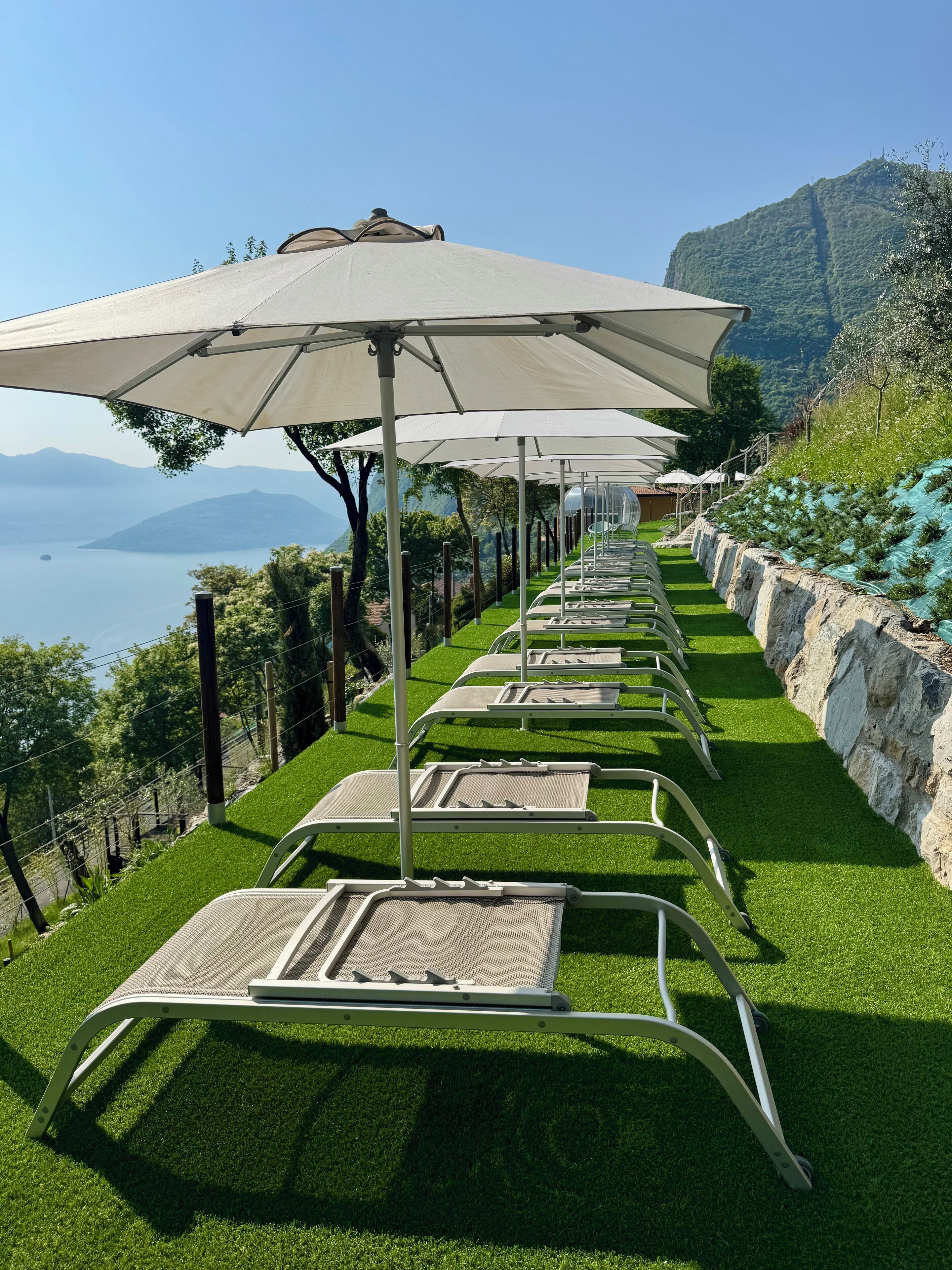 Una hilera de sillones de exterior con sombrillas, dispuestos en una pendiente cubierta de césped con vistas a un lago y montañas al fondo.