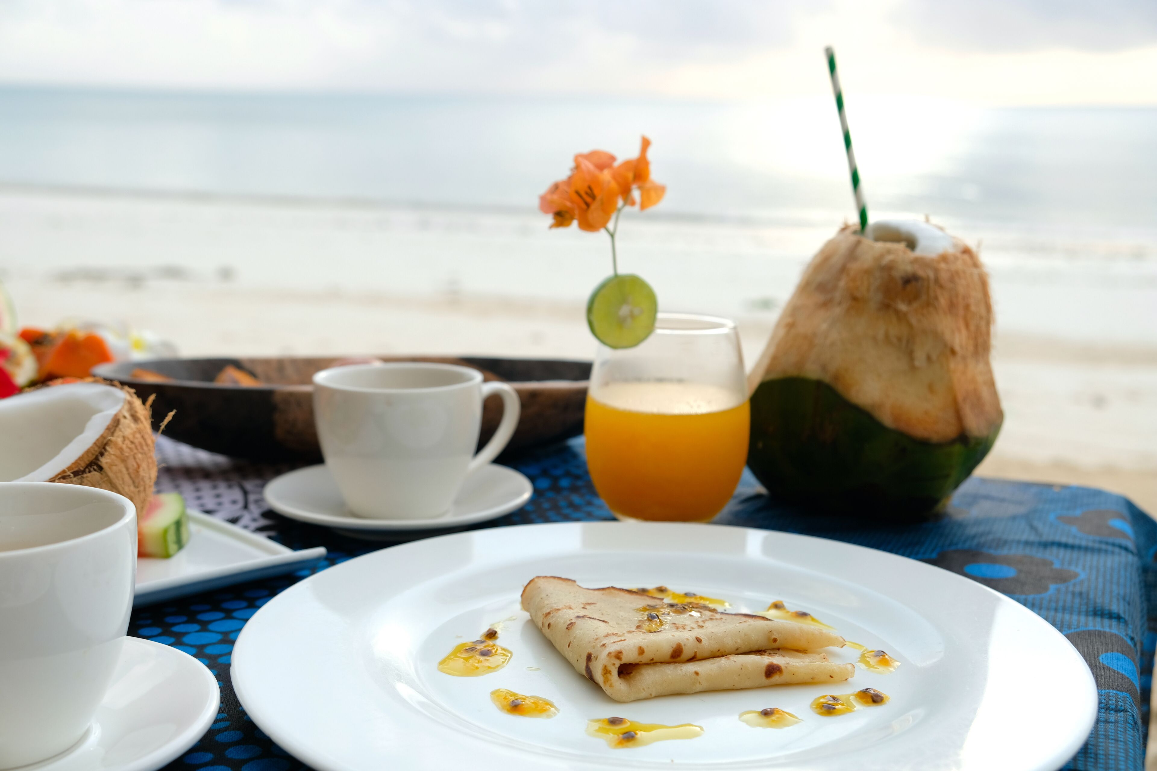 Une table dressée pour le petit-déjeuner, avec une assiette de crêpes, un verre de jus d’orange et une boisson à la noix de coco, le tout placé de manière à profiter de la vue sur la plage.