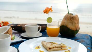 Une table dressée pour le petit-déjeuner, avec une assiette de crêpes, un verre de jus d’orange et une boisson à la noix de coco, le tout placé de manière à profiter de la vue sur la plage.