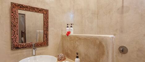 Un bagno con un lavandino bianco, uno specchio con cornice decorata, un soffione doccia a mano e articoli da toilette sul ripiano.