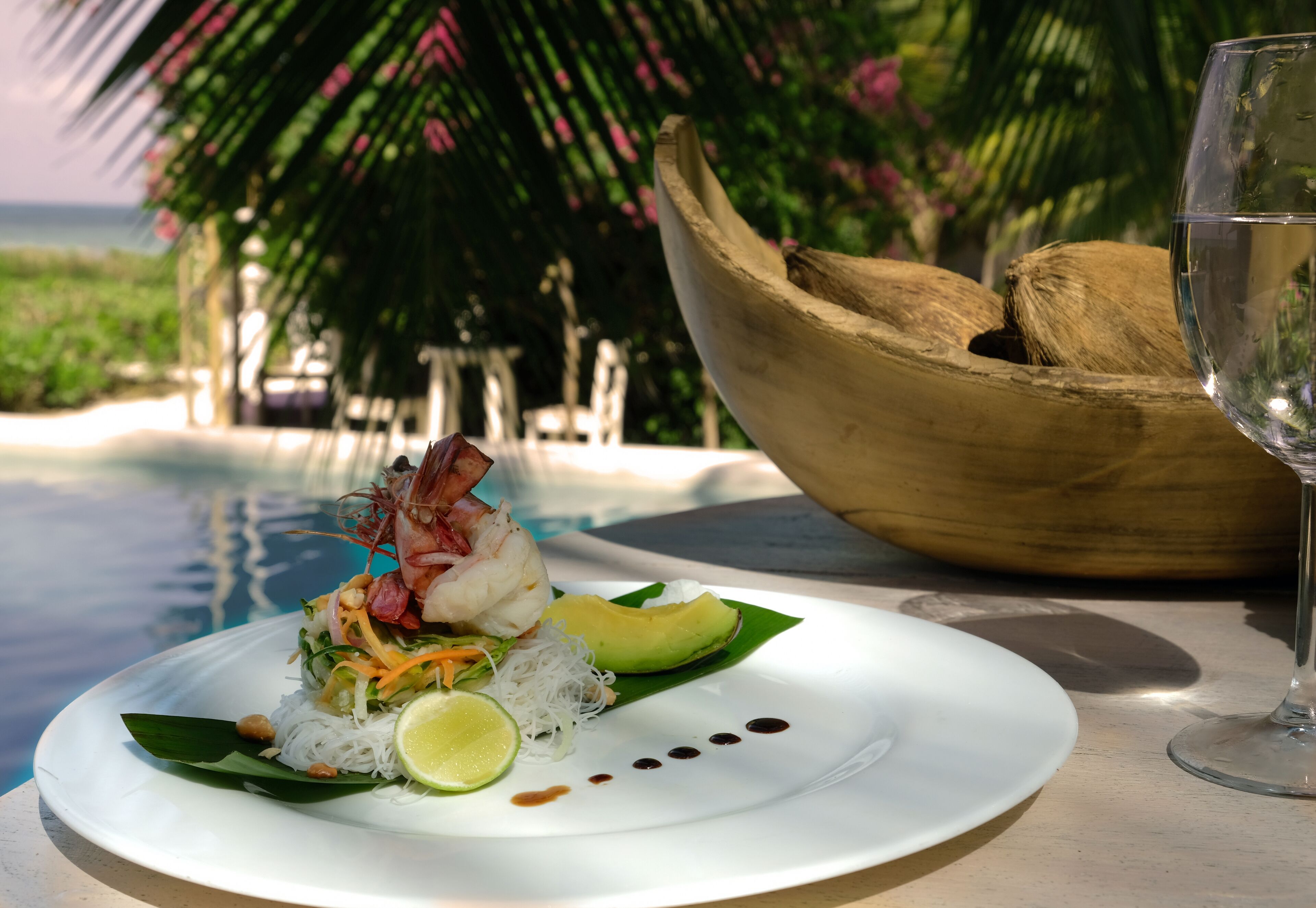 A seafood dish with avocado and lime on a white plate, a glass of water, and a wooden bowl with coconuts.