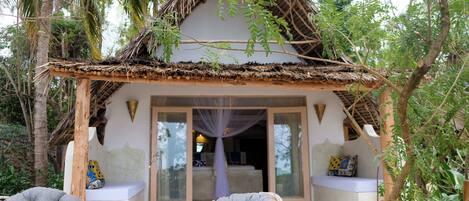 A thatched-roof structure with a glass door entrance, two cushioned chairs, and a bench with cushions.