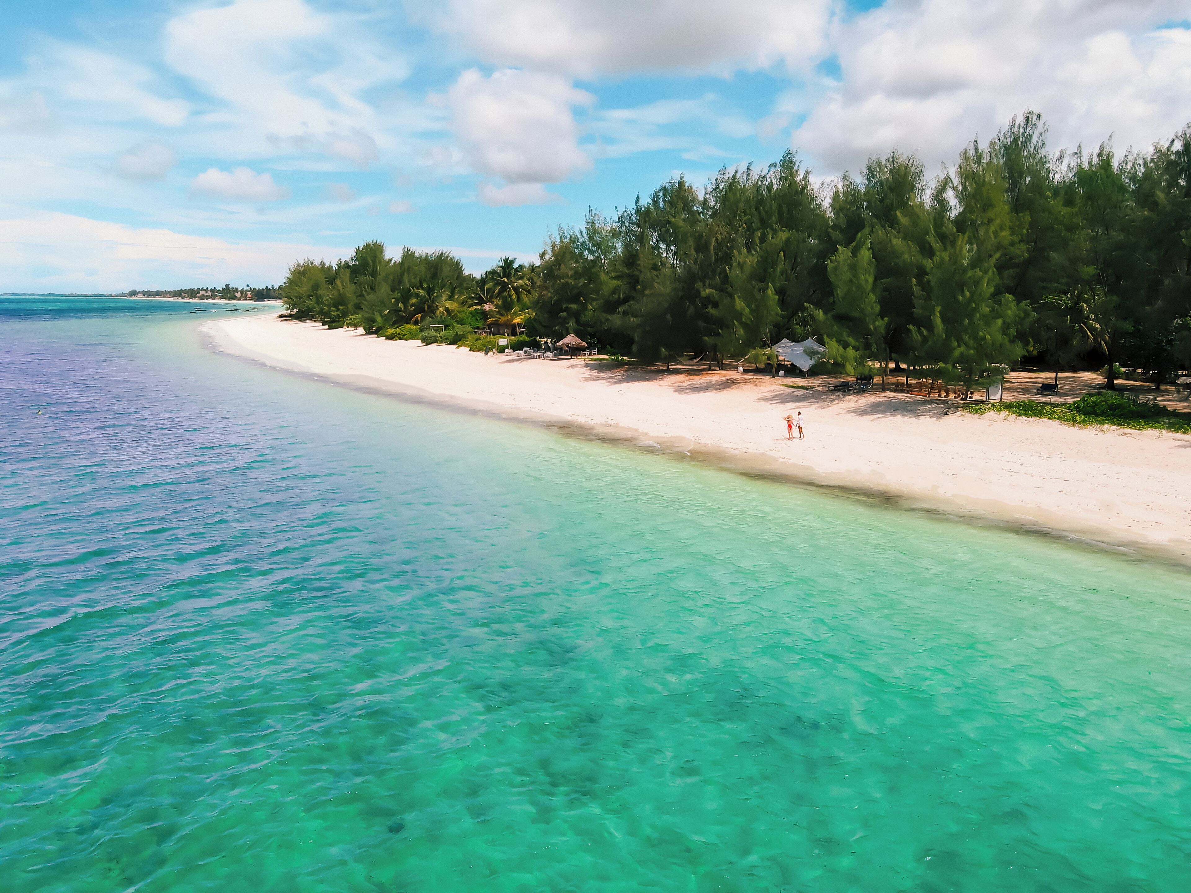 Una playa con agua turquesa cristalina, arena blanca y una hilera de árboles y cabañas al fondo.