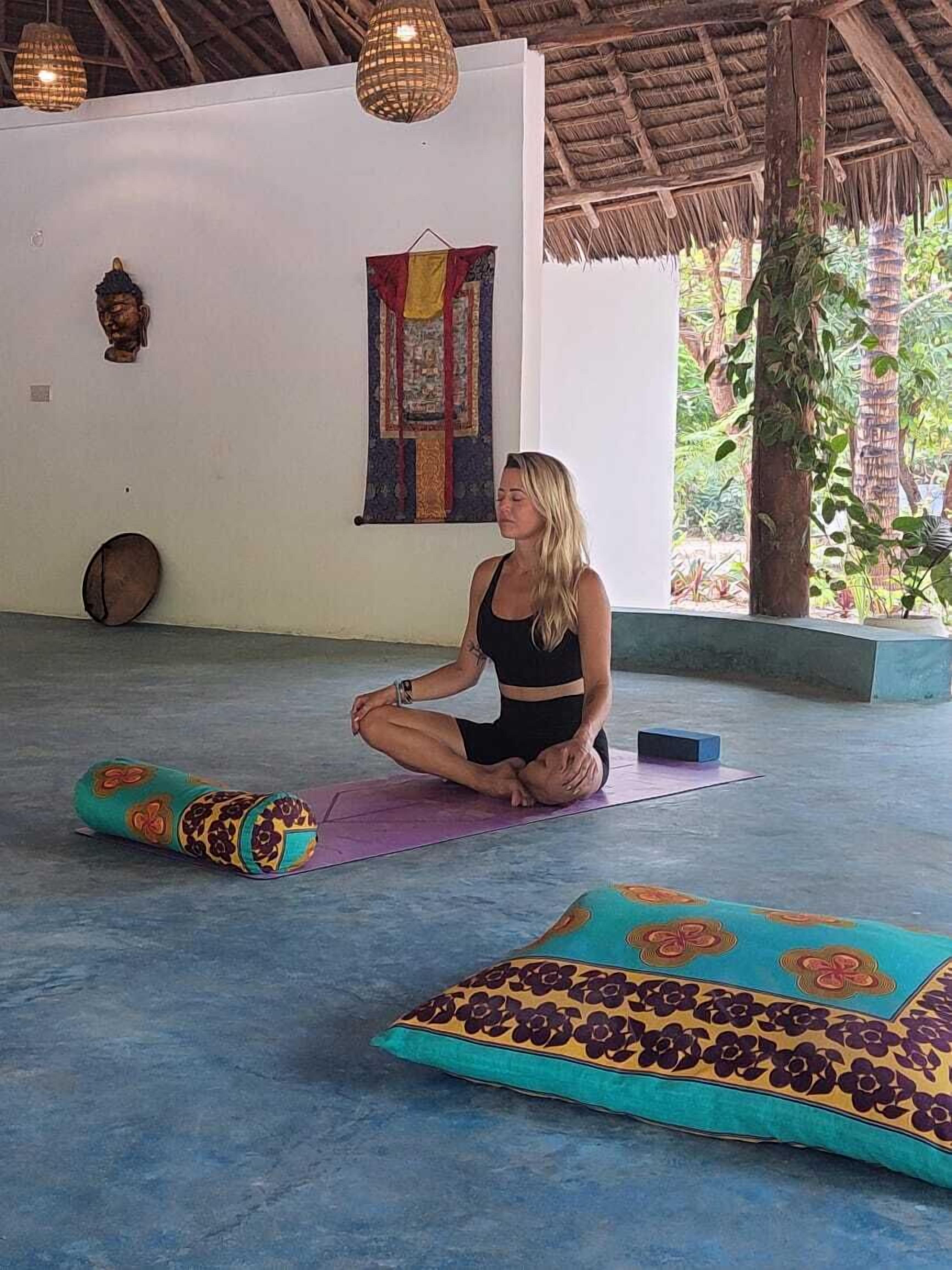 Une personne est assise sur un tapis de yoga dans une pièce dont le toit est fait de chaume, avec une décoration murale et une fenêtre donnant sur un paysage verdoyant.