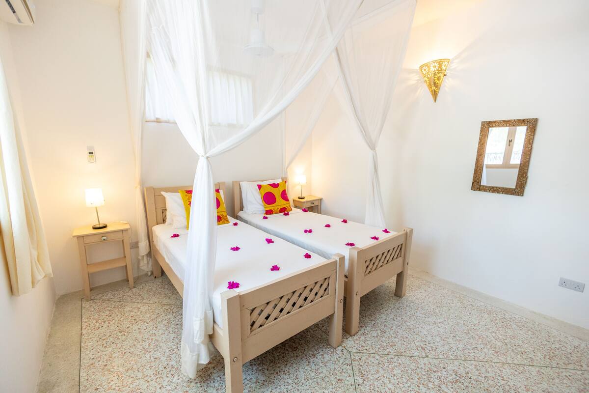 Habitación con dos camas, una cama con dosel, una mesita de noche con lámpara, una luz en la pared y un espejo.