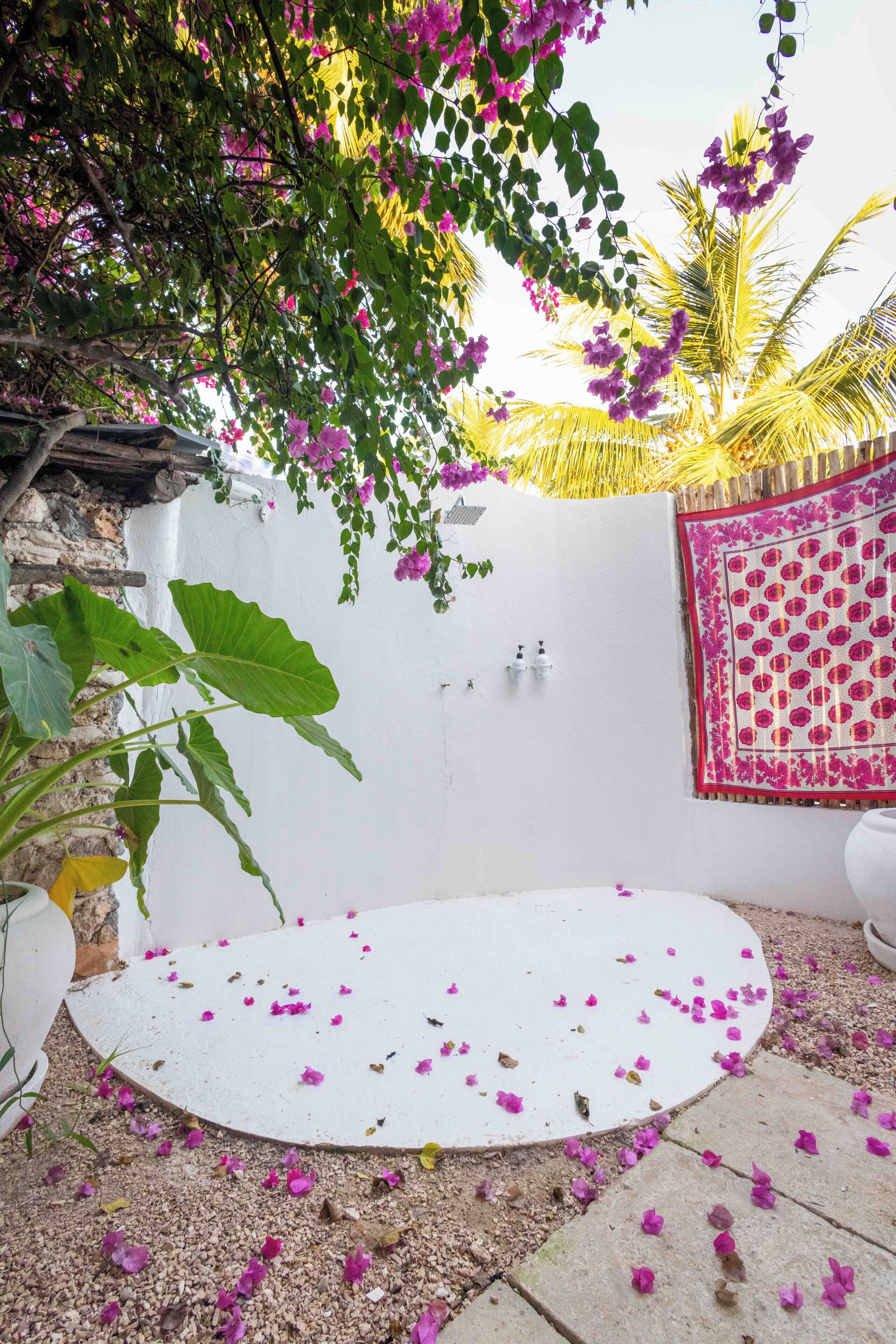 A circular white platform surrounded by scattered flower petals, with a white wall and a decorative pink and red patterned cloth hanging on it.