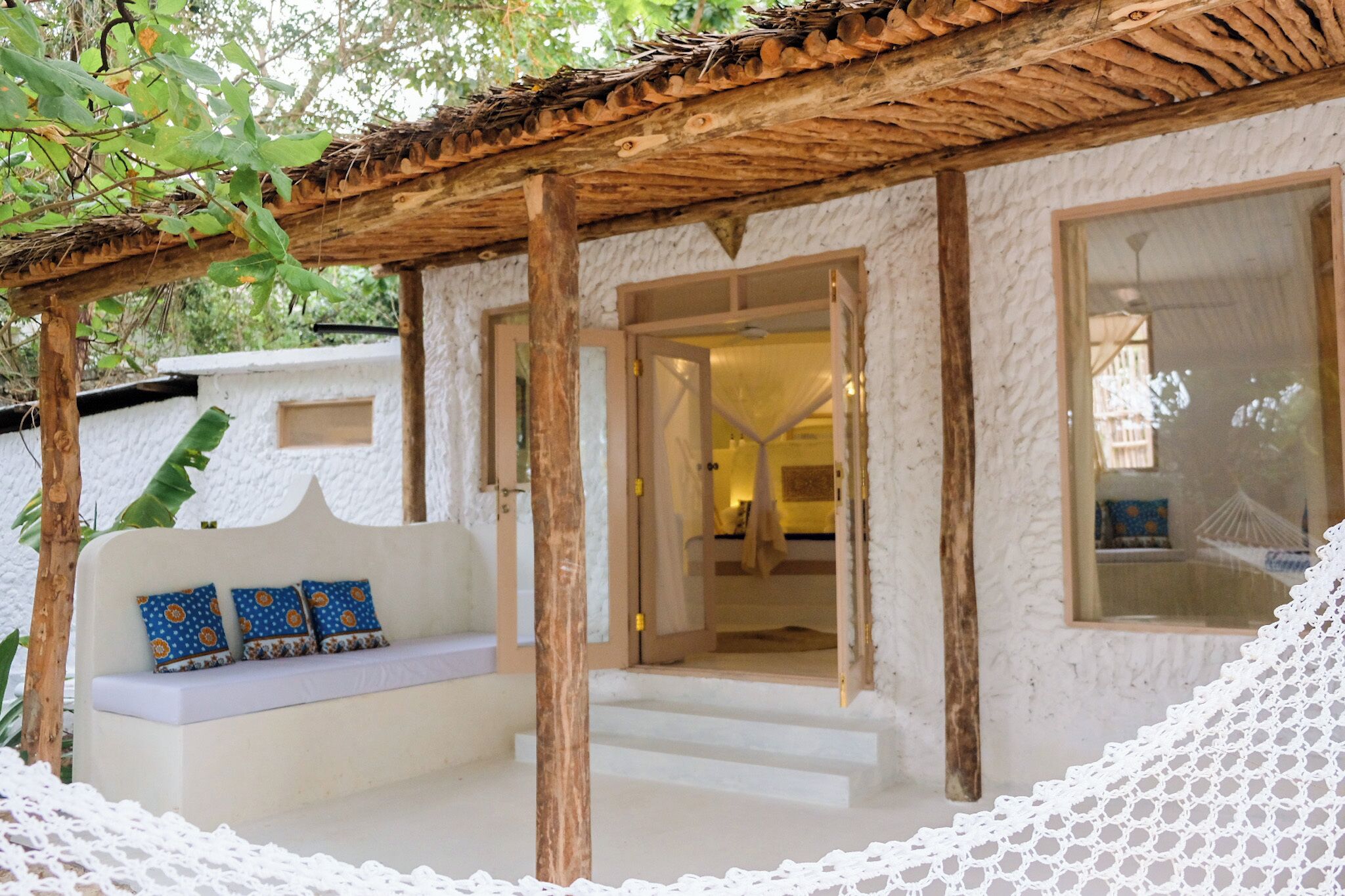 Ein gemütlicher Sitzbereich im Freien mit einer Holz-Pergola, einer weißen Wand mit Struktur und einem großen Fenster.