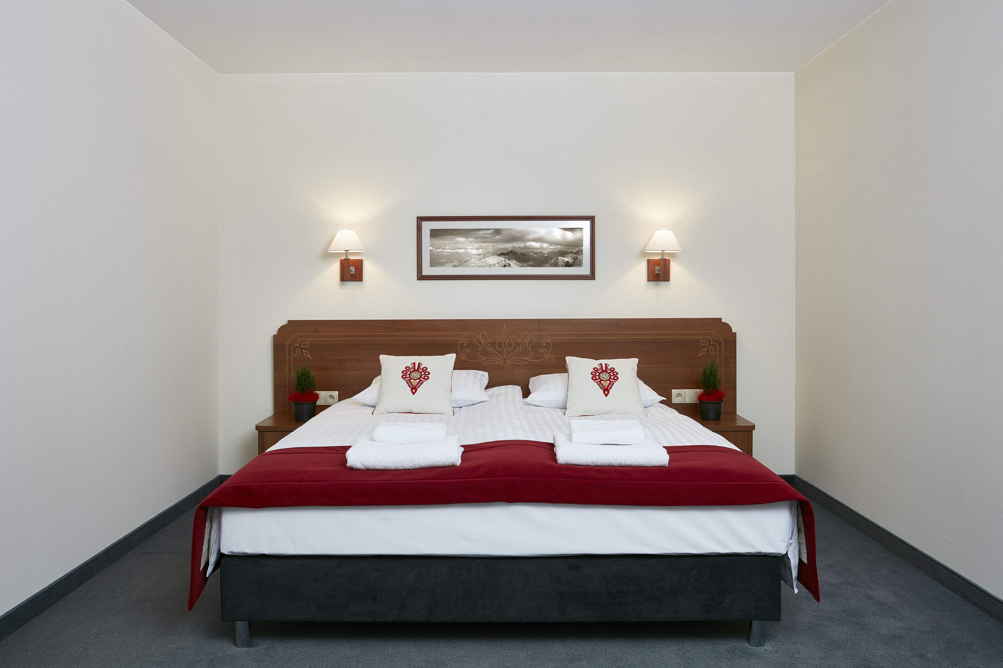 A neatly made bed with white and red linens, two bedside lamps, a framed picture on the wall, and a small potted plant on the nightstand.